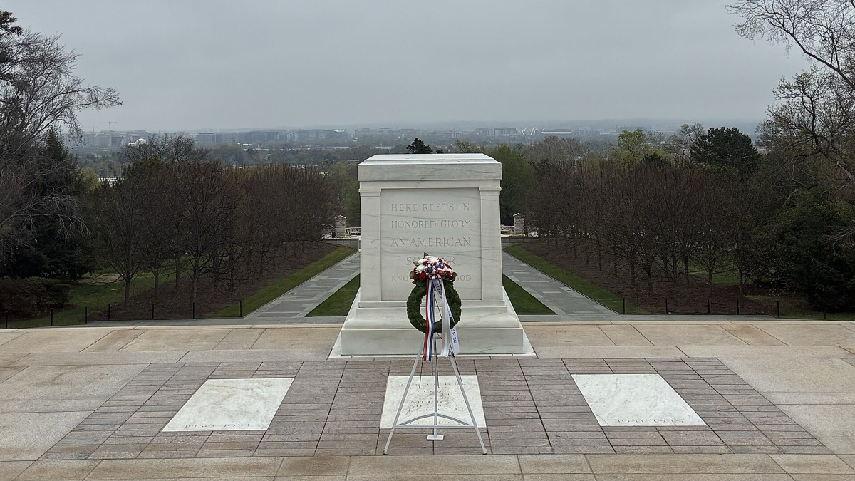 A plain white marble sarcophagus at the head of a wide paved plaza, a wreath on a stand before it, empty trees beyond