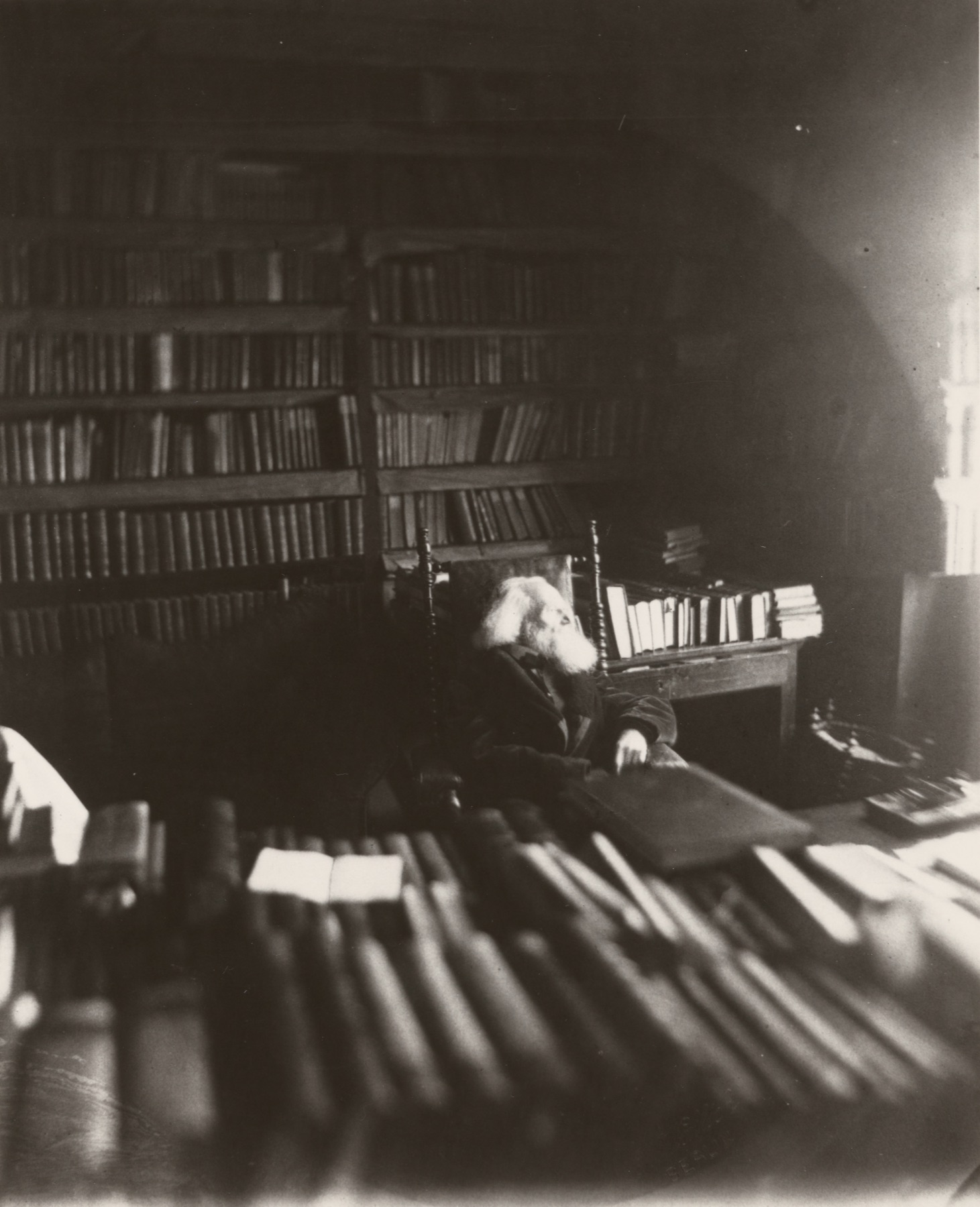 A dim photograph of an old, white-bearded man seated in a high-backed chair, almost lost among floor-to-ceiling shelves of books, with stacks of volumes and an open book piled across the table in the foreground